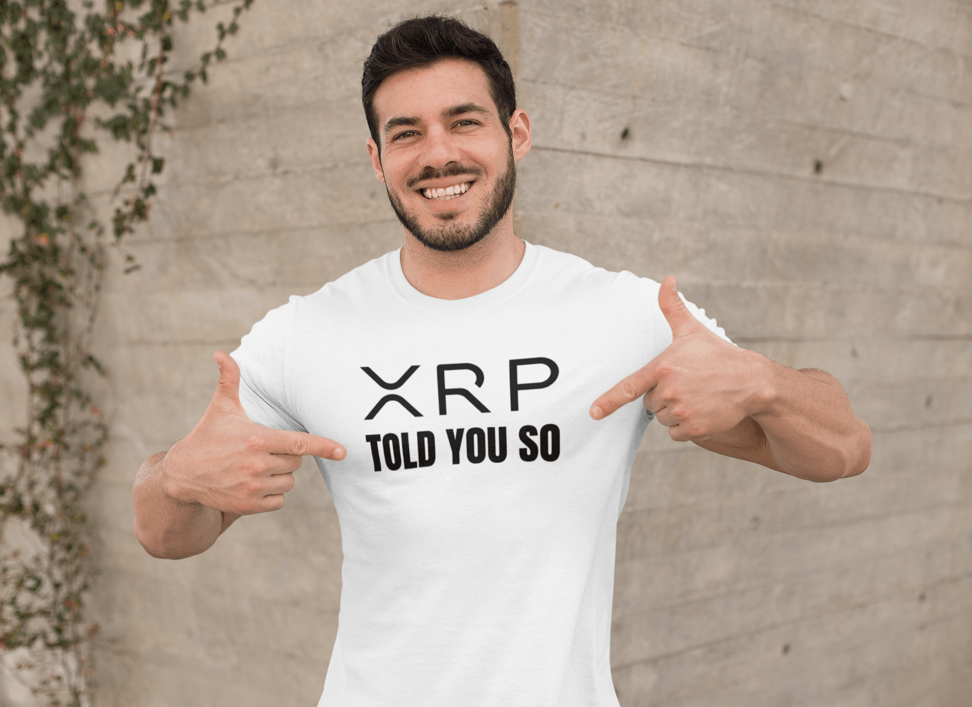 Man wearing a white t-shirt with 'XRP TOLD YOU SO' text, pointing at it, against a concrete wall.